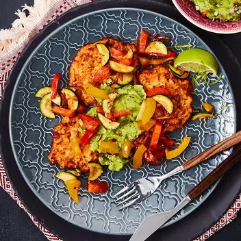 Süßkartoffel-Rösti mit würzigem Fajita-Gemüse aus Paprika, Zucchini und Tomaten, serviert auf einem orientalischen Teller. Beilage: cremige Guacamole in einer Schale, garniert mit frischer Limette und roten Chilischoten. Dekoriert mit einem gemusterten Serviertuch und stilvollem Holz-Besteck.