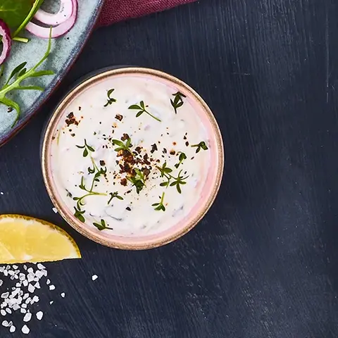 Zwei goldbraun gebratene Gemüsetaler, serviert auf einem Teller mit Kresse-Kräuterquark und einem frischen Salat aus Feldsalat, Radieschen, roten Zwiebeln und einer leichten Vinaigrette. Daneben liegt ein kleines Schälchen mit Quarkdip sowie Zitronenspalten, angerichtet auf einem dunklen Hintergrund mit Besteck auf einer weinroten Stoffserviette.