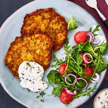 Zwei goldbraun gebratene Gemüsetaler, serviert auf einem Teller mit Kresse-Kräuterquark und einem frischen Salat aus Feldsalat, Radieschen, roten Zwiebeln und einer leichten Vinaigrette. Daneben liegt ein kleines Schälchen mit Quarkdip sowie Zitronenspalten, angerichtet auf einem dunklen Hintergrund mit Besteck auf einer weinroten Stoffserviette.