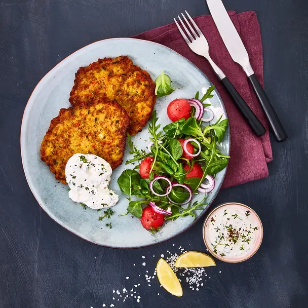 Zwei goldbraun gebratene Gemüsetaler, serviert auf einem Teller mit Kresse-Kräuterquark und einem frischen Salat aus Feldsalat, Radieschen, roten Zwiebeln und einer leichten Vinaigrette. Daneben liegt ein kleines Schälchen mit Quarkdip sowie Zitronenspalten, angerichtet auf einem dunklen Hintergrund mit Besteck auf einer weinroten Stoffserviette.