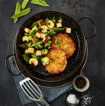 Zwei goldbraune Gemüsetaler, serviert in einer schwarzen Pfanne mit einer Gemüse-Tofu-Mischung aus Zucchini, Brokkoli, Zuckerschoten und knusprig angebratenen Tofuwürfeln, garniert mit dunkler Soße. Neben der Pfanne liegen eine Metall-Bratwender, grobes Meersalz und Pfefferkörner in kleinen Schalen auf einem dunklen Hintergrund.