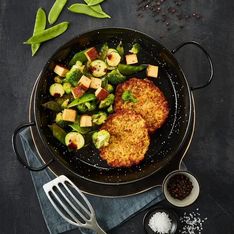 Zwei goldbraune Gemüsetaler, serviert in einer schwarzen Pfanne mit einer Gemüse-Tofu-Mischung aus Zucchini, Brokkoli, Zuckerschoten und knusprig angebratenen Tofuwürfeln, garniert mit dunkler Soße. Neben der Pfanne liegen eine Metall-Bratwender, grobes Meersalz und Pfefferkörner in kleinen Schalen auf einem dunklen Hintergrund.