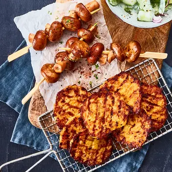 Knusprige Kartoffelgrillis auf einem kleinen Gitter, daneben gegrillte Champignonspieße, serviert auf einem rustikalen Holzbrett mit Backpapier. Als Beilage steht eine Schale mit Gurkensalat, garniert mit roten Zwiebelringen und Dill. Das Arrangement ist auf einem dunklen Untergrund mit blauer Stoffserviette präsentiert.