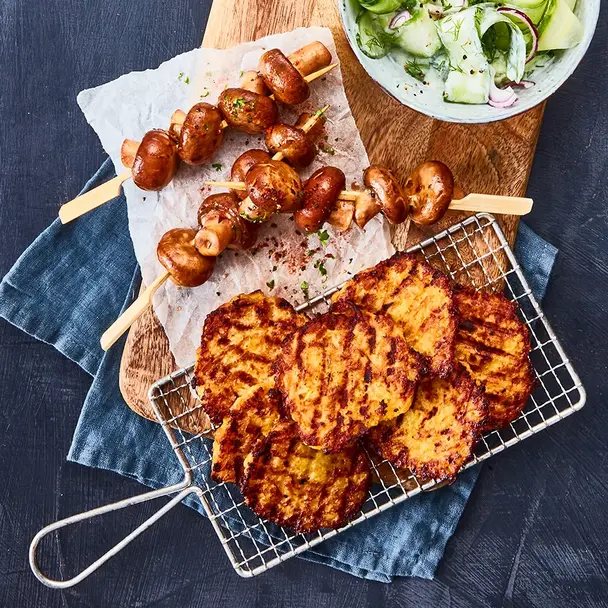 Knusprige Kartoffelgrillis auf einem kleinen Gitter, daneben gegrillte Champignonspieße, serviert auf einem rustikalen Holzbrett mit Backpapier. Als Beilage steht eine Schale mit Gurkensalat, garniert mit roten Zwiebelringen und Dill. Das Arrangement ist auf einem dunklen Untergrund mit blauer Stoffserviette präsentiert.