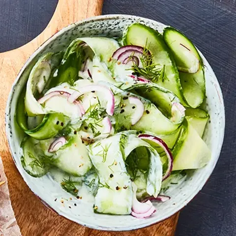 Knusprige Kartoffelgrillis auf einem kleinen Gitter, daneben gegrillte Champignonspieße, serviert auf einem rustikalen Holzbrett mit Backpapier. Als Beilage steht eine Schale mit Gurkensalat, garniert mit roten Zwiebelringen und Dill. Das Arrangement ist auf einem dunklen Untergrund mit blauer Stoffserviette präsentiert.