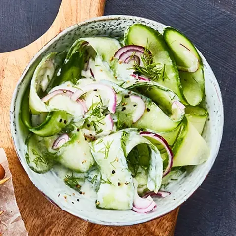 Knusprige Kartoffelgrillis auf einem kleinen Gitter, daneben gegrillte Champignonspieße, serviert auf einem rustikalen Holzbrett mit Backpapier. Als Beilage steht eine Schale mit Gurkensalat, garniert mit roten Zwiebelringen und Dill. Das Arrangement ist auf einem dunklen Untergrund mit blauer Stoffserviette präsentiert.