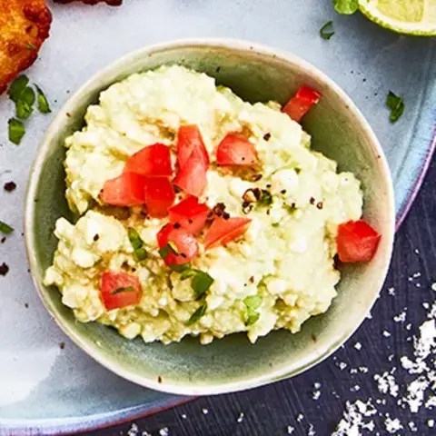 Ein Teller mit drei knusprigen Reibekuchen, serviert mit einer kleinen Schale Avocadocreme, garniert mit gehackten Tomaten und frischen Kräutern. Eine halbe Limette und grobes Meersalz liegen dekorativ daneben. Das Arrangement ist auf einer olivgrünen Stoffserviette präsentiert, begleitet von einer Gabel mit Holzgriff, alles auf einem dunklen Hintergrund.