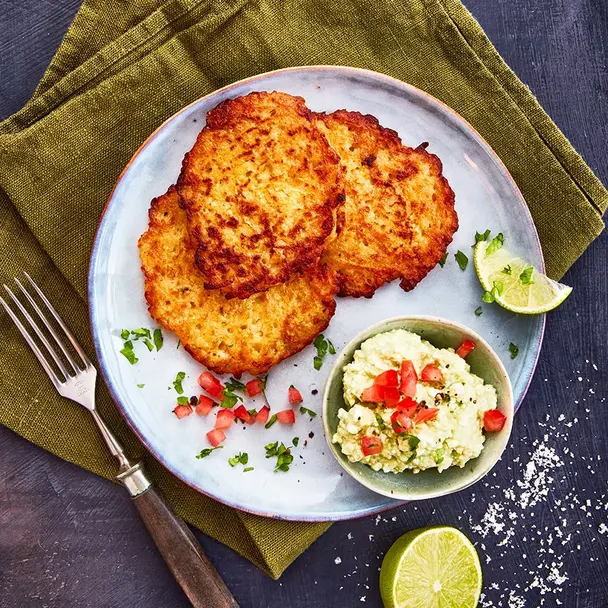 Ein Teller mit drei knusprigen Reibekuchen, serviert mit einer kleinen Schale Avocadocreme, garniert mit gehackten Tomaten und frischen Kräutern. Eine halbe Limette und grobes Meersalz liegen dekorativ daneben. Das Arrangement ist auf einer olivgrünen Stoffserviette präsentiert, begleitet von einer Gabel mit Holzgriff, alles auf einem dunklen Hintergrund.