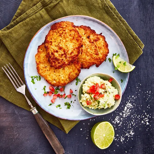 Ein Teller mit drei knusprigen Reibekuchen, serviert mit einer kleinen Schale Avocadocreme, garniert mit gehackten Tomaten und frischen Kräutern. Eine halbe Limette und grobes Meersalz liegen dekorativ daneben. Das Arrangement ist auf einer olivgrünen Stoffserviette präsentiert, begleitet von einer Gabel mit Holzgriff, alles auf einem dunklen Hintergrund.