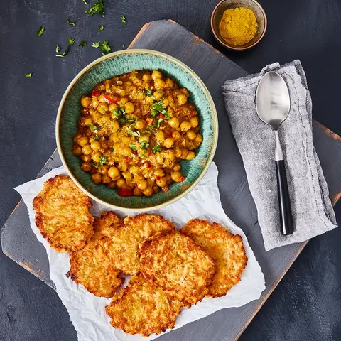 Süßkartoffel-Rösti serviert mit einem aromatischen Kichererbsen-Curry in einer blauen Schale, garniert mit frischen Kräutern. Daneben eine kleine Schale mit Curry-Gewürz und ein dekorativ gefaltetes Stofftuch mit Löffel. Angerichtet auf einer rustikalen Holzplatte.