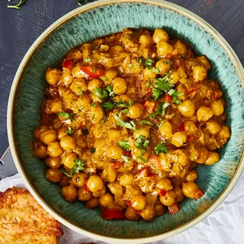 Süßkartoffel-Rösti serviert mit einem aromatischen Kichererbsen-Curry in einer blauen Schale, garniert mit frischen Kräutern. Daneben eine kleine Schale mit Curry-Gewürz und ein dekorativ gefaltetes Stofftuch mit Löffel. Angerichtet auf einer rustikalen Holzplatte.