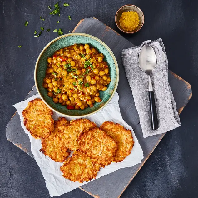 Süßkartoffel-Rösti serviert mit einem aromatischen Kichererbsen-Curry in einer blauen Schale, garniert mit frischen Kräutern. Daneben eine kleine Schale mit Curry-Gewürz und ein dekorativ gefaltetes Stofftuch mit Löffel. Angerichtet auf einer rustikalen Holzplatte.