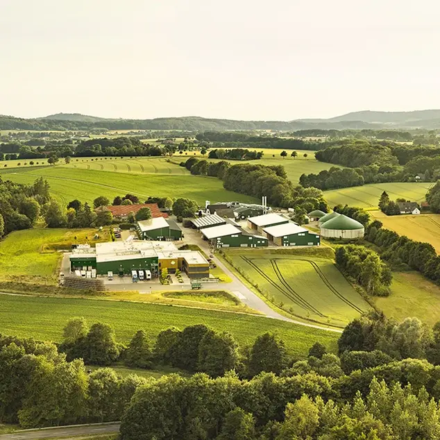 Luftaufnahme des Hofes Pahmeyer und der angeschlossenen Kartoffelmanufaktur, umgeben von weitläufigen Feldern, Wäldern und dem Ravensberger Hügelland im Hintergrund, unter einem leicht bewölkten Himmel. Ein Beispiel für nachhaltige Landwirtschaft in idyllischer Umgebung.