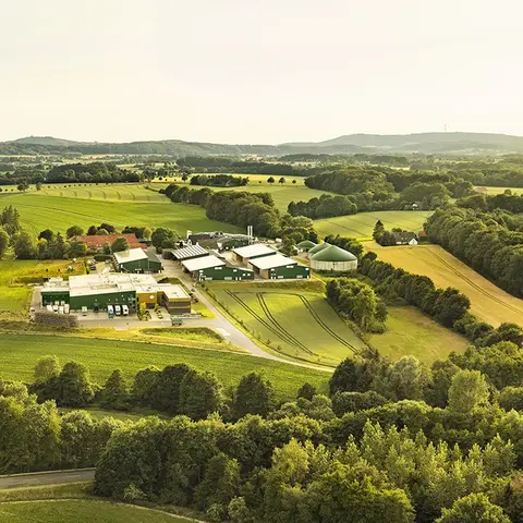 Luftaufnahme des Hofes Pahmeyer und der angeschlossenen Kartoffelmanufaktur, umgeben von weitläufigen Feldern, Wäldern und dem Ravensberger Hügelland im Hintergrund, unter einem leicht bewölkten Himmel. Ein Beispiel für nachhaltige Landwirtschaft in idyllischer Umgebung.