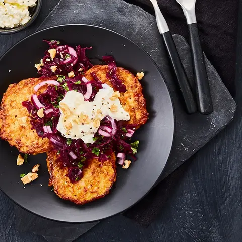 Drei knusprige Süßkartoffel-Rösti auf einem schwarzen Teller, garniert mit würzigem Rotkrautsalat, zerbröckeltem Fetakäse und gehackten Nüssen. Die dunkle Schieferplatte und das stilvolle Besteck runden die edle Präsentation ab. Eine kleine Schale mit cremigem Feta-Dip mit Olivenöl ergänzt das Gericht. 