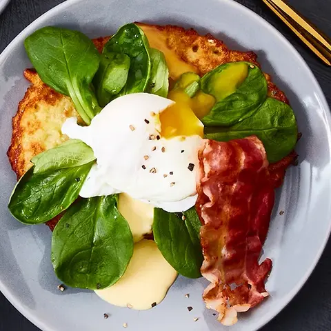 Drei Teller mit Reibekuchen, belegt mit Feldsalat, einem pochierten Ei, knusprigem Bacon und einer cremigen Soße, garniert mit frisch gemahlenem Pfeffer. Die Teller sind mit goldfarbenem Besteck arrangiert. Daneben stehen kleine Schalen mit Pfefferkörnern und zusätzlicher Soße, alles auf einem dunklen Hintergrund präsentiert.