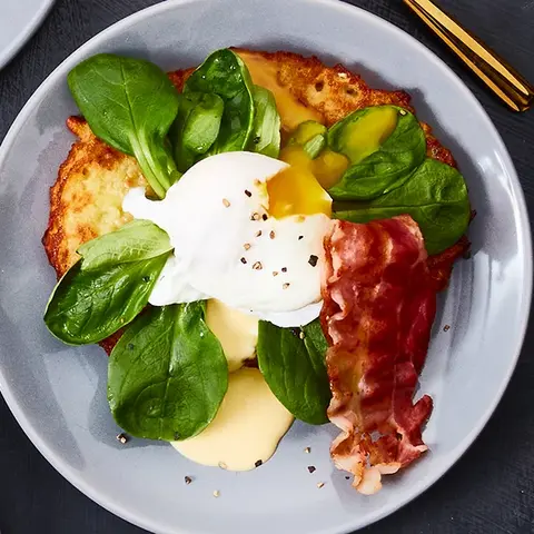 Drei Teller mit Reibekuchen, belegt mit Feldsalat, einem pochierten Ei, knusprigem Bacon und einer cremigen Soße, garniert mit frisch gemahlenem Pfeffer. Die Teller sind mit goldfarbenem Besteck arrangiert. Daneben stehen kleine Schalen mit Pfefferkörnern und zusätzlicher Soße, alles auf einem dunklen Hintergrund präsentiert.