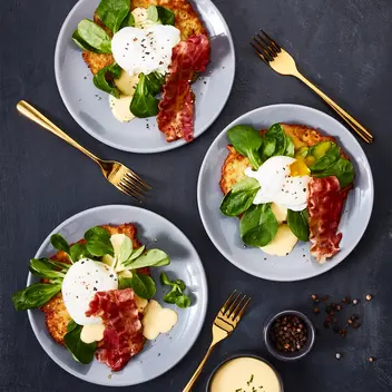 Drei Teller mit Reibekuchen, belegt mit Feldsalat, einem pochierten Ei, knusprigem Bacon und einer cremigen Soße, garniert mit frisch gemahlenem Pfeffer. Die Teller sind mit goldfarbenem Besteck arrangiert. Daneben stehen kleine Schalen mit Pfefferkörnern und zusätzlicher Soße, alles auf einem dunklen Hintergrund präsentiert.