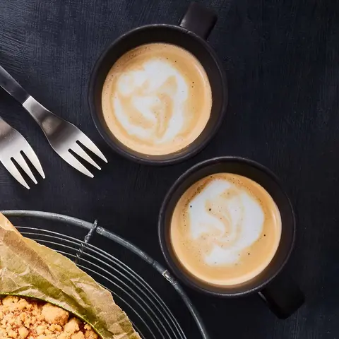 Ein frisch gebackener Apfelkuchen mit Streuseln, serviert auf Backpapier und einem Gitter. Daneben liegen Apfelscheiben, zwei Kaffeetassen mit Latte Art und Gabeln auf einem dunklen Hintergrund.