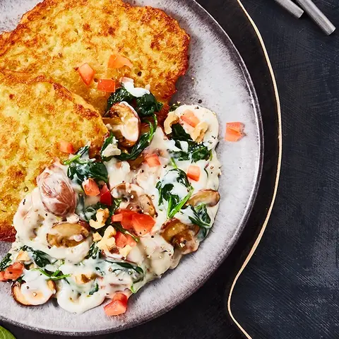 Reibekuchen serviert mit einer cremigen Gorgonzola-Sauce, Spinat und Champignons, dekoriert mit Walnüssen auf dunklem Untergrund.