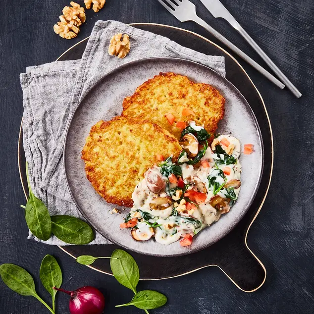 Reibekuchen serviert mit einer cremigen Gorgonzola-Sauce, Spinat und Champignons, dekoriert mit Walnüssen auf dunklem Untergrund.