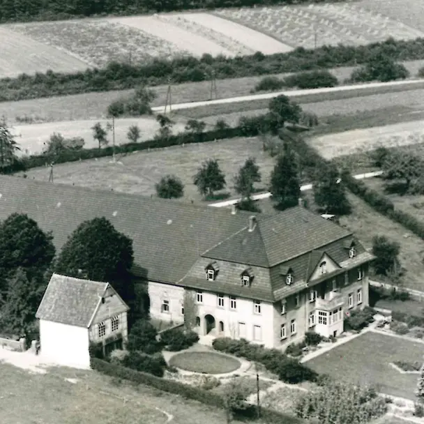 Historische Schwarz-Weiß-Luftaufnahme des Hofes Pahmeyer. Das Bild zeigt eine klassische Hofanlage mit mehreren Gebäuden, darunter ein großes Wohnhaus und angrenzende Scheunen, eingebettet in eine ländliche Umgebung mit Feldern und Wiesen. Die Gebäude sind von Bäumen und gepflegten Gärten umgeben, und Wege führen durch die Landschaft.