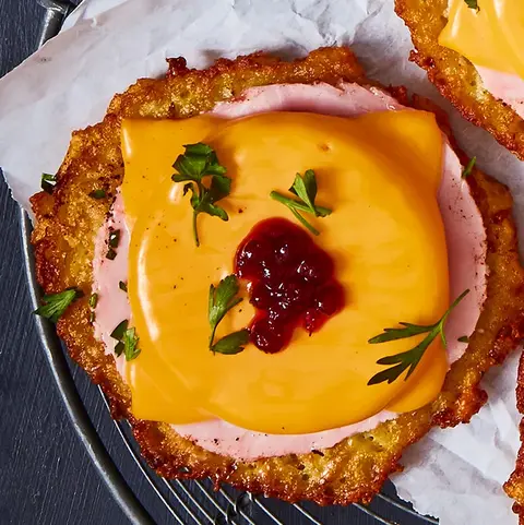 Drei Reibekuchen, belegt im Stil eines Toast Hawaii mit einer Scheibe Schinken, geschmolzenem Käse und einem Klecks Preiselbeermarmelade, dekoriert mit frischen Kräutern. Die Reibekuchen sind auf einem Gitter mit Backpapier angerichtet, ein weiterer liegt auf einem separaten Teller mit einer Gabel. Daneben steht eine kleine Schale mit Preiselbeeren in Soße, alles präsentiert auf einem dunklen Hintergrund mit einer blauen Stoffserviette.