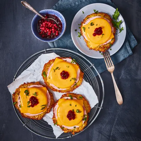 Drei Reibekuchen, belegt im Stil eines Toast Hawaii mit einer Scheibe Schinken, geschmolzenem Käse und einem Klecks Preiselbeermarmelade, dekoriert mit frischen Kräutern. Die Reibekuchen sind auf einem Gitter mit Backpapier angerichtet, ein weiterer liegt auf einem separaten Teller mit einer Gabel. Daneben steht eine kleine Schale mit Preiselbeeren in Soße, alles präsentiert auf einem dunklen Hintergrund mit einer blauen Stoffserviette.