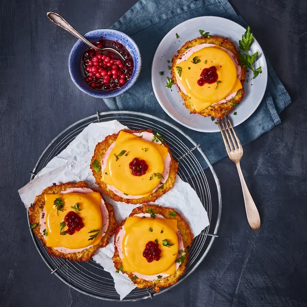 Drei Reibekuchen, belegt im Stil eines Toast Hawaii mit einer Scheibe Schinken, geschmolzenem Käse und einem Klecks Preiselbeermarmelade, dekoriert mit frischen Kräutern. Die Reibekuchen sind auf einem Gitter mit Backpapier angerichtet, ein weiterer liegt auf einem separaten Teller mit einer Gabel. Daneben steht eine kleine Schale mit Preiselbeeren in Soße, alles präsentiert auf einem dunklen Hintergrund mit einer blauen Stoffserviette.