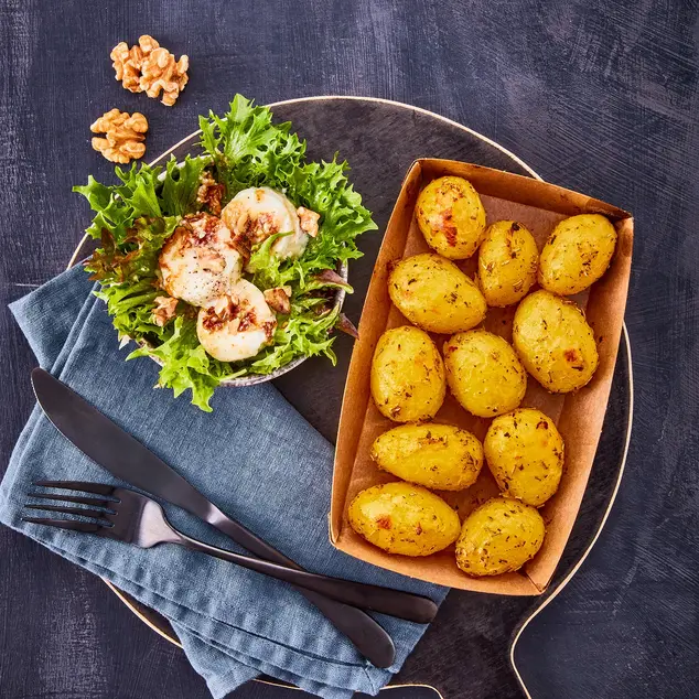 Rosmarinkartoffeln in einer Backschale, serviert mit einem frischen grünen Salat, getoppt mit karamellisiertem Honig-Ziegenkäse und Walnüssen, angerichtet auf einem dunklen Servierbrett mit blauer Stoffserviette und schwarzem Besteck.