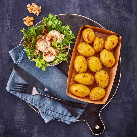 Rosmarinkartoffeln in einer Backschale, serviert mit einem frischen grünen Salat, getoppt mit karamellisiertem Honig-Ziegenkäse und Walnüssen, angerichtet auf einem dunklen Servierbrett mit blauer Stoffserviette und schwarzem Besteck.