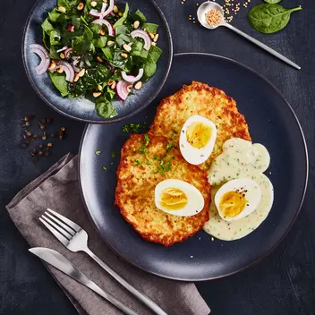 Ein Teller mit zwei Reibekuchen, serviert mit halbierten hartgekochten Eiern und einer Kräutersoße, garniert mit frischen Kräutern. Daneben steht eine Schale mit frischem Spinatsalat, garniert mit roten Zwiebelringen und gerösteten Körnern. as Arrangement wird von frischen Spinatblättern und einem Löffel mit Sesam auf einem dunklen Hintergrund ergänzt, begleitet von Besteck auf einer braunen Stoffserviette.