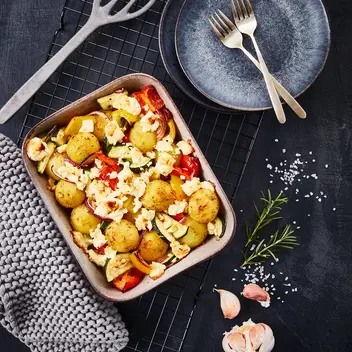 Rosmarinkartoffeln mit Ofengemüse wie Zucchini, Paprika und roten Zwiebeln, überbacken mit zerbröseltem Feta-Käse, serviert in einer Backform auf einem schwarzen Gitter. Das Gericht wird von einer grauen Topflappen-Dekoration, Rosmarinzweig, Knoblauchzehen und grobem Meersalz ergänzt. Zwei moderne Teller mit goldenen Gabeln stehen bereit.