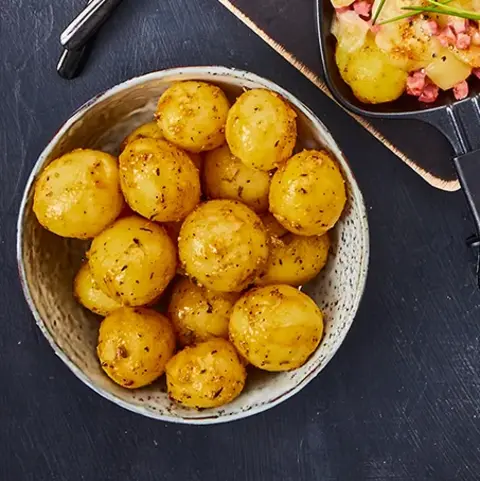 ine ansprechende Präsentation von Raclette-Gerichten in drei Pfännchen auf einem dunklen Untergrund. Die Pfännchen enthalten Variationen aus Kartoffeln, Gemüse, Käse und Speck. Begleitend stehen kleine Schälchen mit Rosmarinkartoffeln, Essiggurken, Silberzwiebeln und einem frischen Tomatensalat. Zwei schwarze Gabeln sind dekorativ platziert, ergänzt durch einen Zweig Rosmarin. Ein köstliches Arrangement für Raclette-Liebhaber.