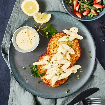 Ein Teller mit zwei knusprigen Reibekuchen, belegt mit Spargelragout und garniert mit frischen Kräutern. Daneben ein Schälchen mit heller Soße und zwei Zitronenscheiben. Im Hintergrund eine weitere Schale mit einem frischen Salat aus Rucola und Erdbeeren. Das Arrangement ist auf einer grünen Stoffserviette präsentiert, begleitet von schwarzem Besteck und angerichtet auf einem dunklen Untergrund.