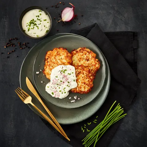 Knusprige Schinken-Rösti serviert mit cremiger Schnittlauchcreme und gehackten roten Zwiebeln, angerichtet auf einem dunkelgrünen Teller. Dekoriert mit frischen Schnittlauchhalmen und goldfarbenem Besteck, auf einem schwarzen Stoffservietten-Hintergrund.