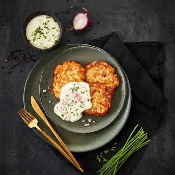 Knusprige Schinken-Rösti serviert mit cremiger Schnittlauchcreme und gehackten roten Zwiebeln, angerichtet auf einem dunkelgrünen Teller. Dekoriert mit frischen Schnittlauchhalmen und goldfarbenem Besteck, auf einem schwarzen Stoffservietten-Hintergrund.