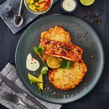 in Teller mit zwei Reibekuchen, belegt mit einem Stück glasiertem Teriyaki-Lachs, dekoriert mit einer gegrillten Limettenscheibe und frischem Koriander. Als Beilage gibt es Avocadostücke und einen Klecks würziger Creme, garniert mit Chiliflocken. Im Hintergrund ein Glas mit Mango-Salsa, daneben schwarze Sesamsamen auf einem Löffel, eine halbe Limette und rote Chilischoten. Alles arrangiert auf einem dunklen Untergrund mit grauer Stoffserviette und Besteck.