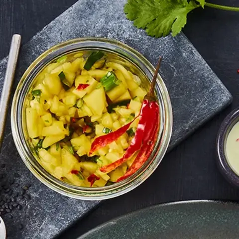 in Teller mit zwei Reibekuchen, belegt mit einem Stück glasiertem Teriyaki-Lachs, dekoriert mit einer gegrillten Limettenscheibe und frischem Koriander. Als Beilage gibt es Avocadostücke und einen Klecks würziger Creme, garniert mit Chiliflocken. Im Hintergrund ein Glas mit Mango-Salsa, daneben schwarze Sesamsamen auf einem Löffel, eine halbe Limette und rote Chilischoten. Alles arrangiert auf einem dunklen Untergrund mit grauer Stoffserviette und Besteck.