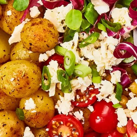 osmarinkartoffeln in einer Bowl mit frischem Salat aus Tomaten, Kichererbsen, Radicchio, Feldsalat und Frischkäse, serviert auf einem Holzbrett. Das Gericht ist mit frischem Rosmarin, Salz und einem grünen Stoffserviette dekoriert. Eine Gabel und ein Messer aus Edelstahl liegen neben der Bowl.