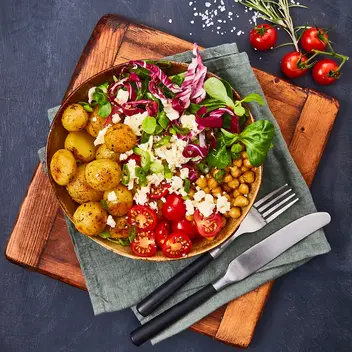 osmarinkartoffeln in einer Bowl mit frischem Salat aus Tomaten, Kichererbsen, Radicchio, Feldsalat und Frischkäse, serviert auf einem Holzbrett. Das Gericht ist mit frischem Rosmarin, Salz und einem grünen Stoffserviette dekoriert. Eine Gabel und ein Messer aus Edelstahl liegen neben der Bowl.