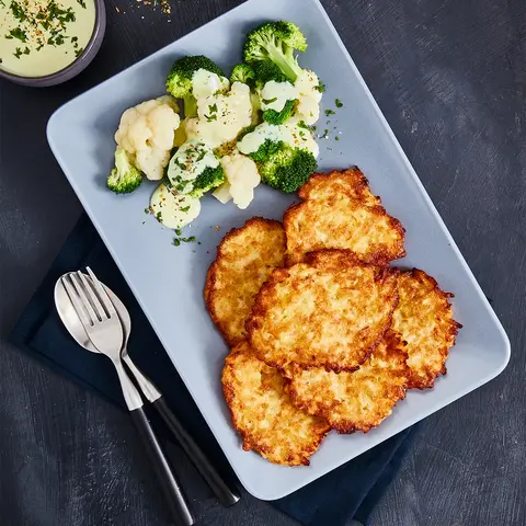 Krosse Rösti, serviert mit gedünstetem Blumenkohl und Brokkoli, garniert mit cremiger Sauce und Kräutern auf einem blauen Teller. Im Hintergrund frische Gewürze und eine passende Sauce im Schälchen.