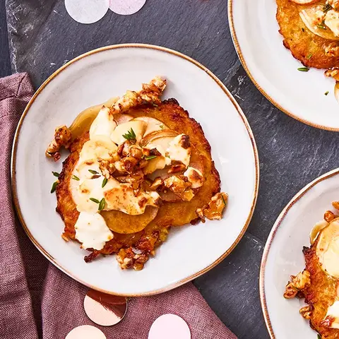 Vier Teller mit Mini-Reibekuchen, belegt mit Birnenscheiben, geschmolzenem Ziegenkäse und karamellisierten Walnüssen, garniert mit frischen Kräutern. Das Arrangement ist auf einer dunklen Schieferplatte angerichtet, dekoriert mit roséfarbenen Konfetti und einer violetten Stoffserviette. Daneben liegen roséfarbene Gabeln, alles präsentiert auf einem dunklen Hintergrund.