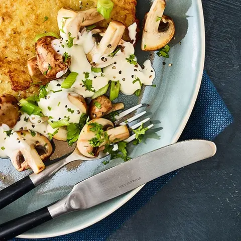 Knusprige Reibekuchen mit einer herzhaften Pilz-Schmand-Sauce, angerichtet auf einem blauen Teller mit Besteck.