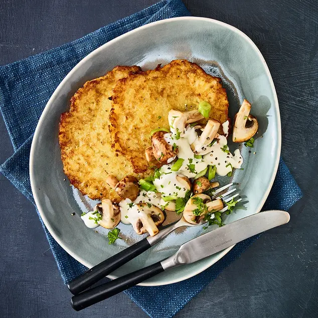 Knusprige Reibekuchen mit einer herzhaften Pilz-Schmand-Sauce, angerichtet auf einem blauen Teller mit Besteck.