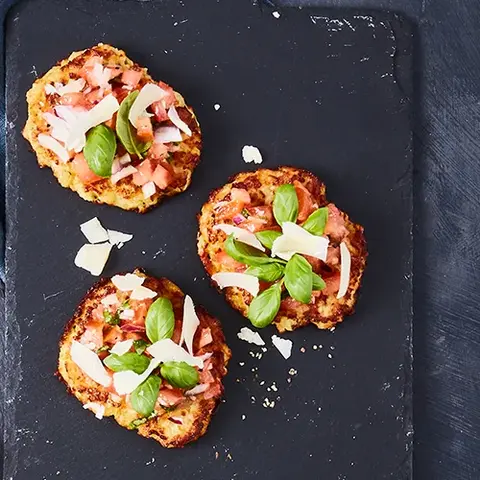 Eine kreative Variation von Reibekuchen als Mini-Bruschetta auf einer dunklen Servierplatte angerichtet. Die knusprigen Kartoffelpuffer sind mit einer frischen Mischung aus Tomaten, Zwiebeln, Basilikum und Parmesan belegt. Einzelne Portionen sind zusätzlich mit Erdbeeren und roten Zwiebeln verfeinert. 