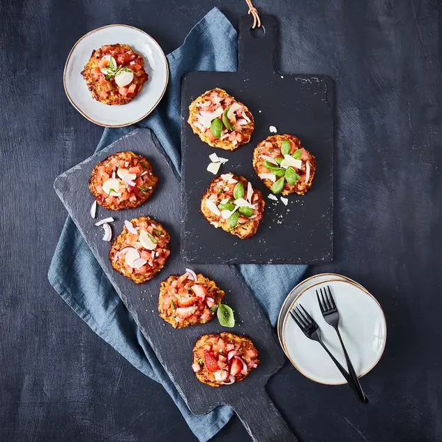 Eine kreative Variation von Reibekuchen als Mini-Bruschetta auf einer dunklen Servierplatte angerichtet. Die knusprigen Kartoffelpuffer sind mit einer frischen Mischung aus Tomaten, Zwiebeln, Basilikum und Parmesan belegt. Einzelne Portionen sind zusätzlich mit Erdbeeren und roten Zwiebeln verfeinert. 