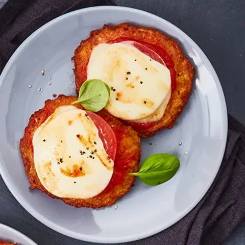 Drei Teller mit Reibekuchen-Minis im Pizza-Margarita-Stil, belegt mit Tomatensoße, geschmolzenem Käse und frischen Basilikumblättern. Die Teller sind dekorativ auf dunklen Servietten und einem Holzbrett arrangiert, begleitet von schwarzen Gabeln. Daneben eine Schale mit grobem Meersalz und verstreuten Basilikumblättern, alles präsentiert auf einem dunklen Untergrund.