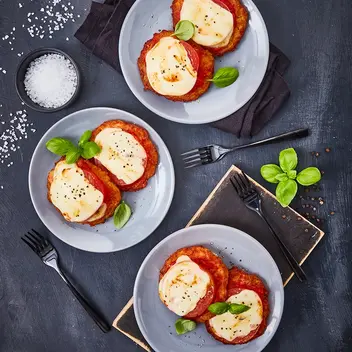 Drei Teller mit Reibekuchen-Minis im Pizza-Margarita-Stil, belegt mit Tomatensoße, geschmolzenem Käse und frischen Basilikumblättern. Die Teller sind dekorativ auf dunklen Servietten und einem Holzbrett arrangiert, begleitet von schwarzen Gabeln. Daneben eine Schale mit grobem Meersalz und verstreuten Basilikumblättern, alles präsentiert auf einem dunklen Untergrund.