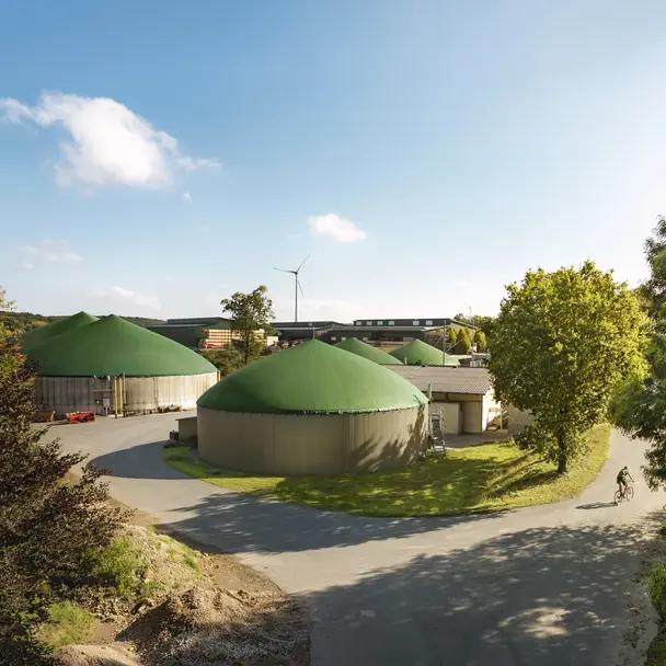 Biogasanlage mit mehreren grünen Silos, umgeben von Bäumen und blühender Natur, unter einem sonnigen Himmel. Eine Person fährt mit dem Fahrrad auf der Straße, die das nachhaltige und umweltfreundliche Konzept unterstreicht.